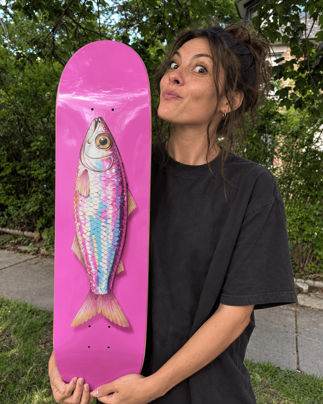 Fish wall art on a hot-pink skateboard deck: an iridescent fish with pink, turquoise and silver scales held upright outdoors in front of green trees